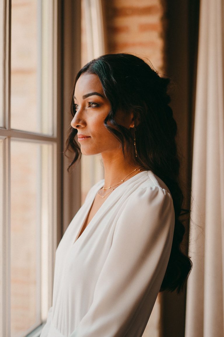 Mariée regardant par la fenêtre, profil gauche, portant une robe blanche avec des manches longues et des bijoux discrets.