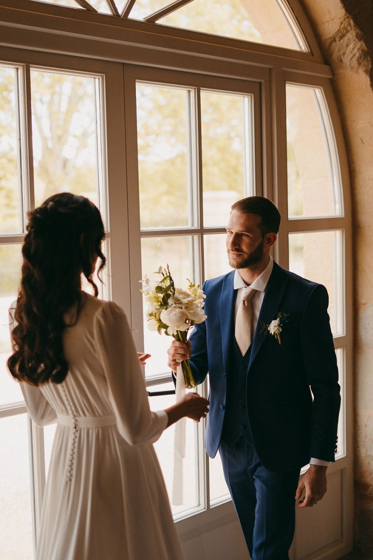 Marié en costume bleu foncé offrant un bouquet à la mariée devant une grande fenêtre.