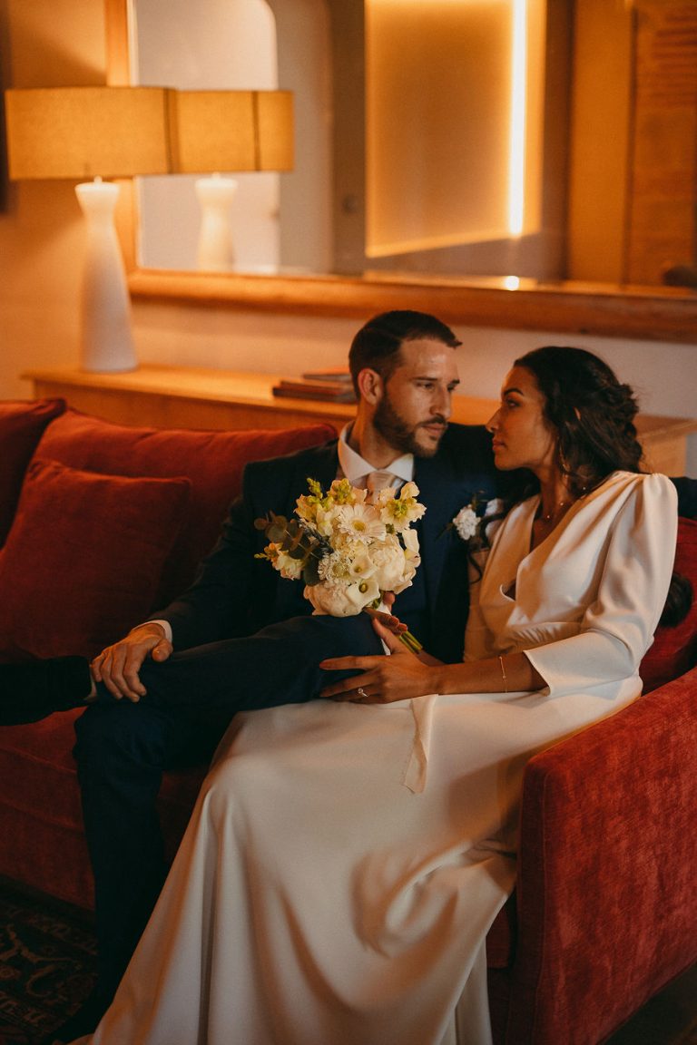 Mariés posant ensemble avec le marié en costume bleu et la mariée en robe blanche, assis sur un canapé rouge, tenant un bouquet de fleurs.