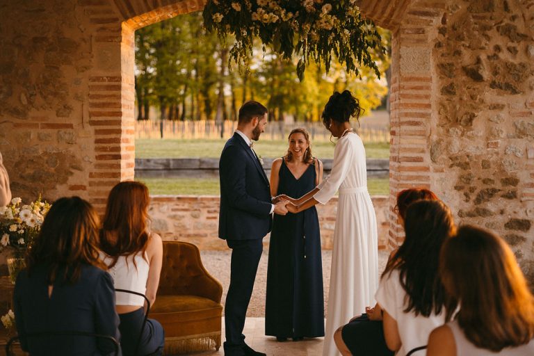 La mariée et le marié se tiennent la main pendant la cérémonie, avec une officiante et des témoins souriants.