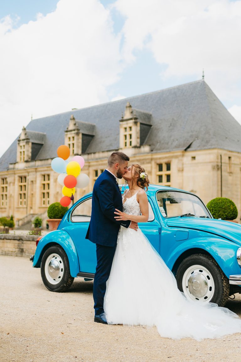 Couple de mariés embrassant devant une voiture bleue décorée de ballons colorés.