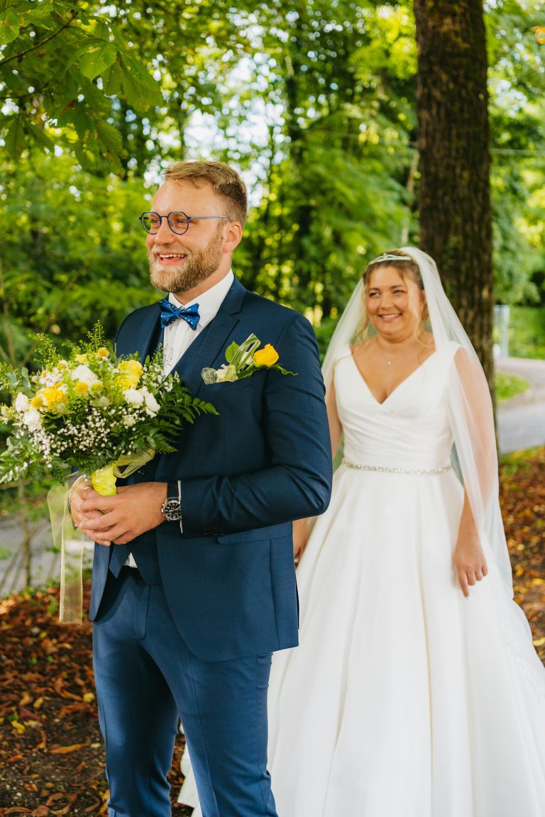 Premier regard entre les mariés, le marié ému tenant un bouquet, avec la mariée souriante en arrière-plan.