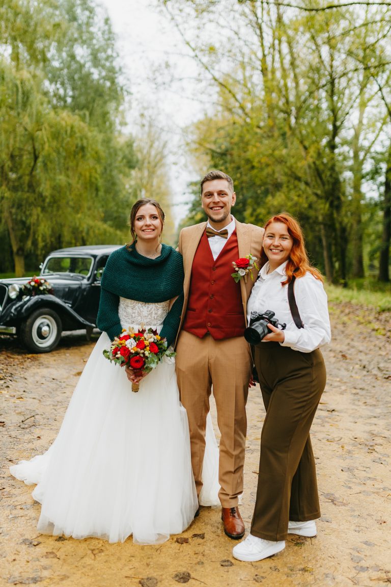 Laura Vergne, photographe mariage, posant avec un couple de mariés dans un décor automnal devant une voiture ancienne.