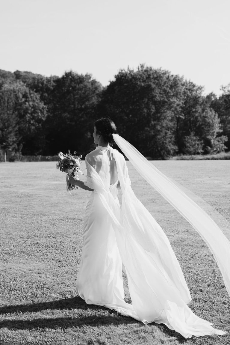 Une mariée portant une robe fluide avec une cape aérienne et un voile, tenant un bouquet dans un champ ensoleillé.