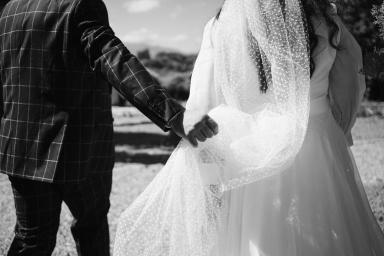 Un marié tenant la main de la mariée, dont le voile en plumetis léger flotte sous le soleil.