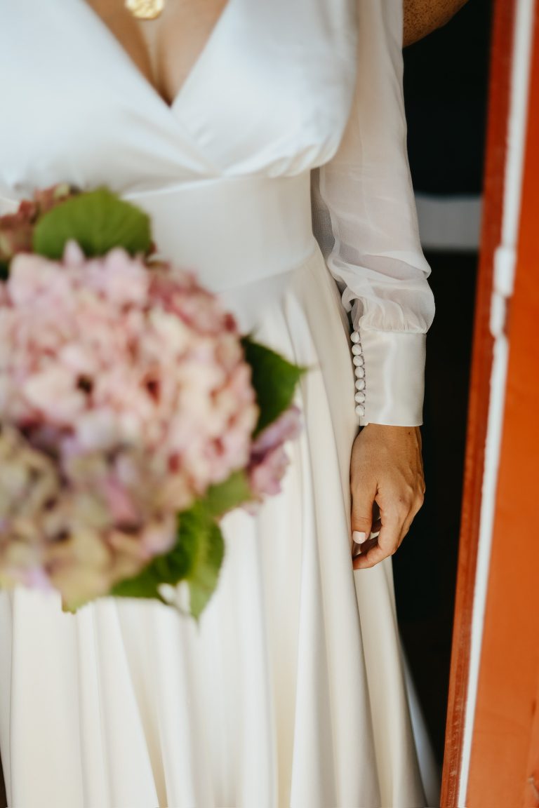 Une mariée portant une robe en satin à manches longues avec des boutons délicats, tenant un bouquet d’hortensias.