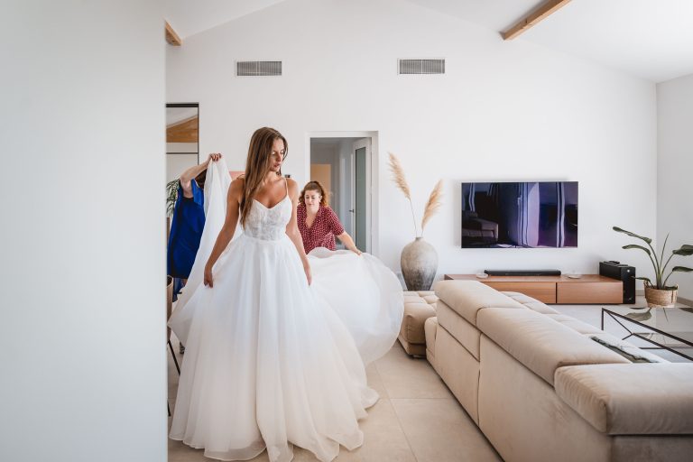 Une mariée en robe princesse ajustée par deux personnes, dans un intérieur moderne et lumineux