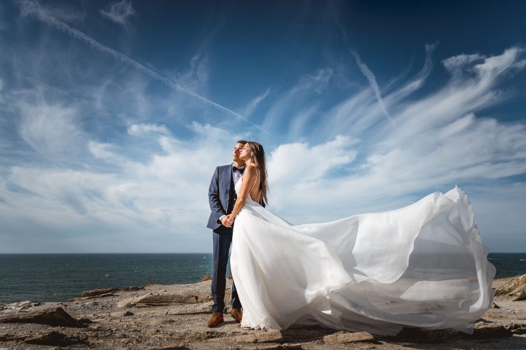 Un couple de mariés sur un bord de mer, la mariée porte une robe fluide en organza qui s'envole avec le vent.