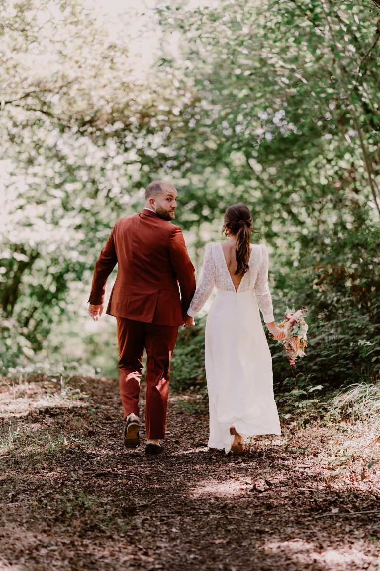 Un couple de mariés marchant main dans la main dans un cadre forestier, la mariée porte une robe bohème en dentelle