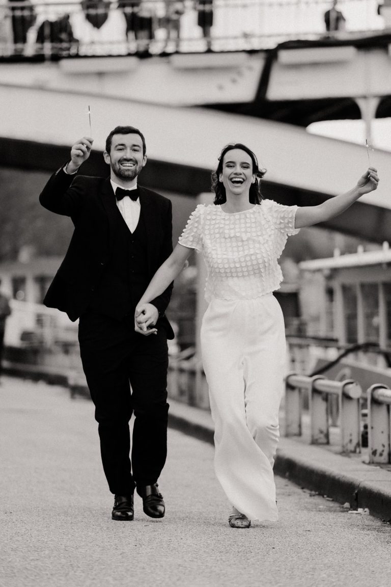 Un couple de mariés souriant et marchant main dans la main, la mariée porte une élégante combinaison-pantalon blanche.