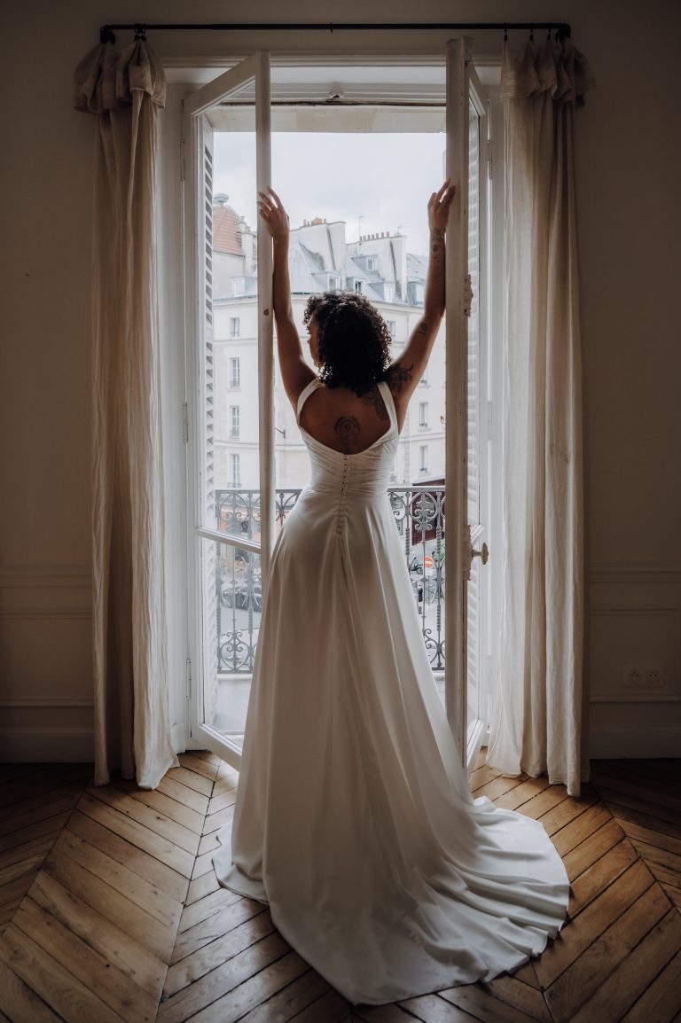 Robe de mariée sur-mesure avec une traîne fluide, vue de dos, dans un appartement parisien.