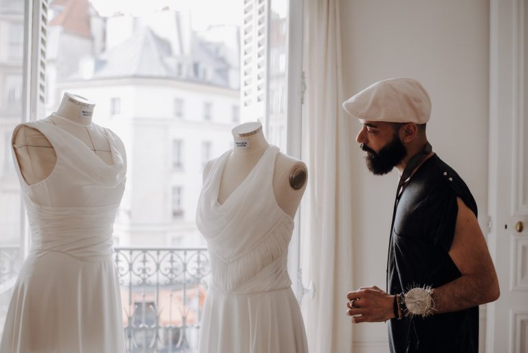 Créateur de robes de mariée sur-mesure observant deux mannequins avec des robes blanches drapées dans un atelier lumineux.