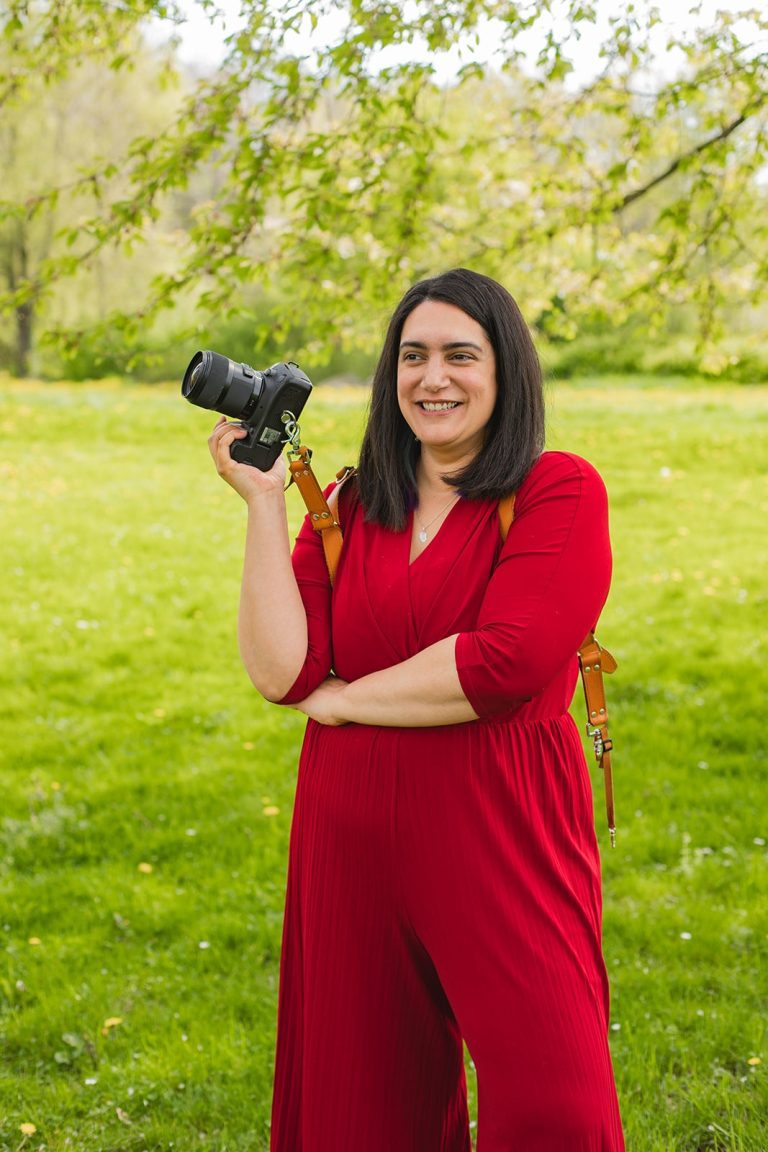 Photographe professionnelle souriante en robe rouge dans un parc verdoyant, tenant un appareil photo reflex.