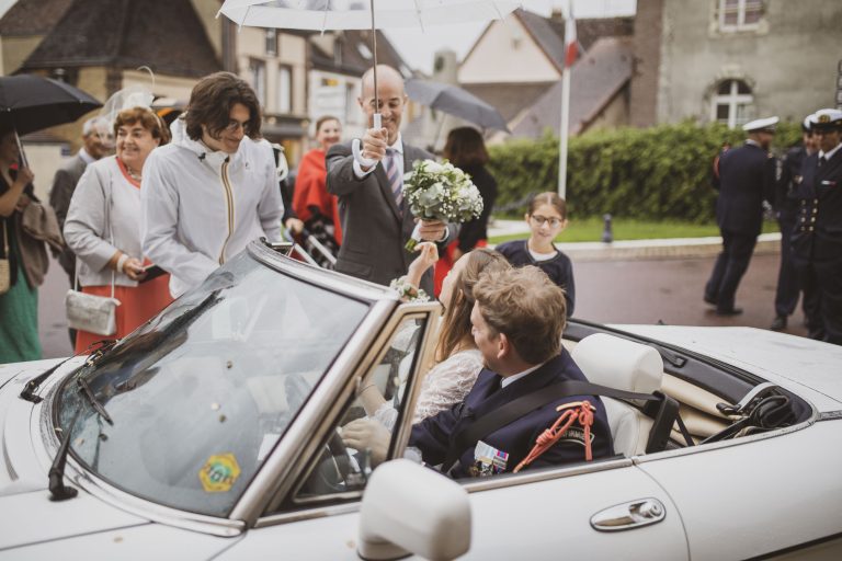 Départ des mariés en décapotable capturé sous la pluie avec des invités spontanés.