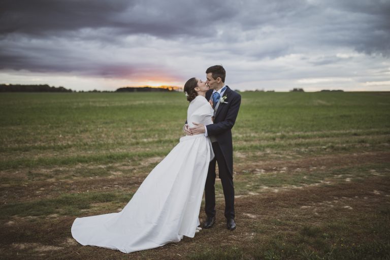 Couple de mariés élégants s'embrassant dans un champ au coucher du soleil, style classique de mariage.