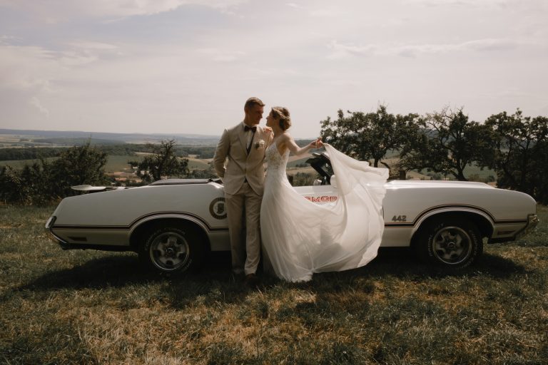 Couple de mariés posant avec une voiture vintage lors d’un mariage tendance 2025.