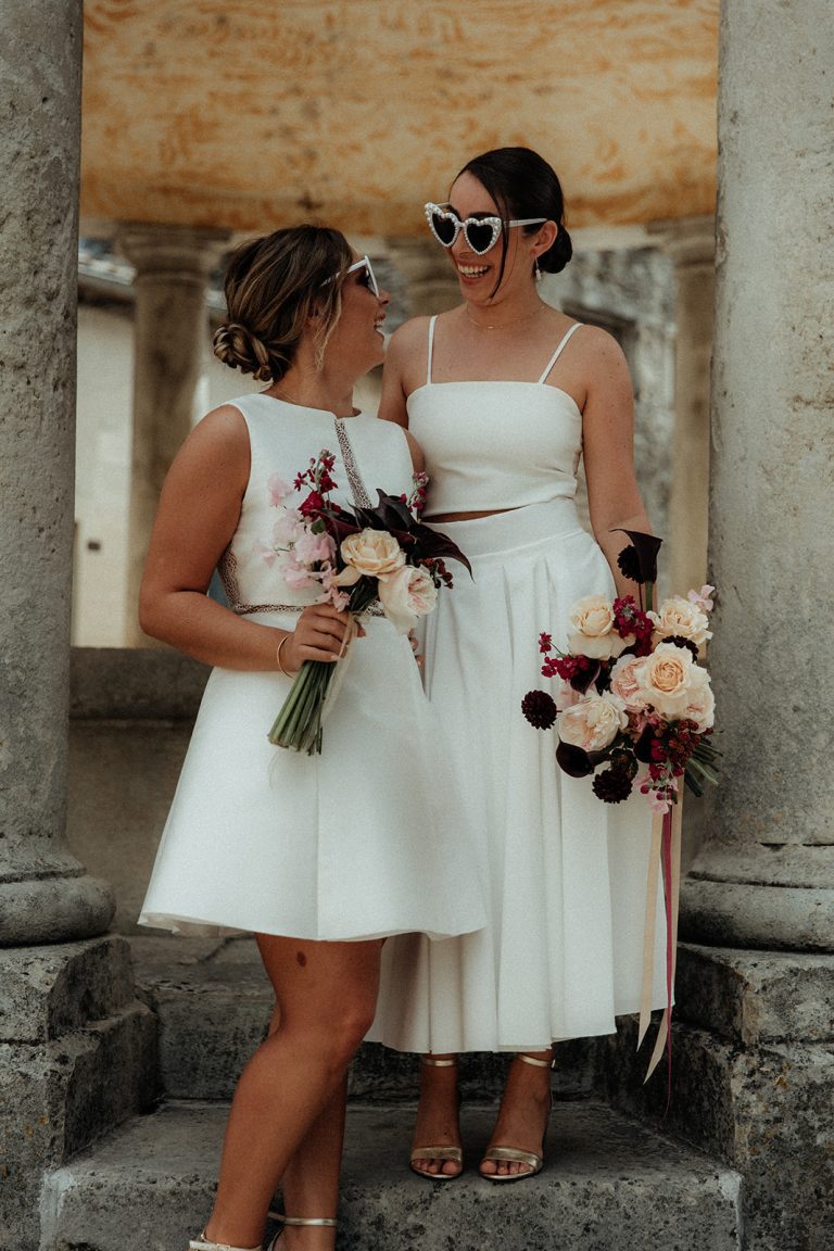 Deux mariées en tenues blanches modernes, souriantes, tenant des bouquets fleuris, avec des lunettes de soleil originales.